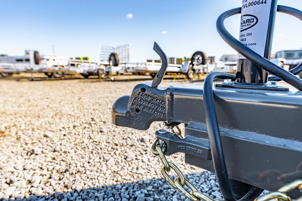 Close-up of a trailer hitch and safety chain on a boat trailer in a gravel lot, with other trailers in the background.