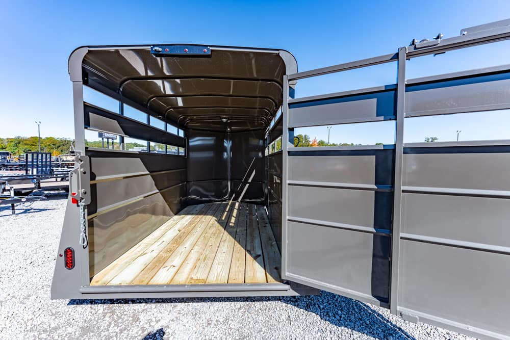 Empty metal livestock trailer interior with a wooden floor and open rear doors on a gravel lot under a blue sky