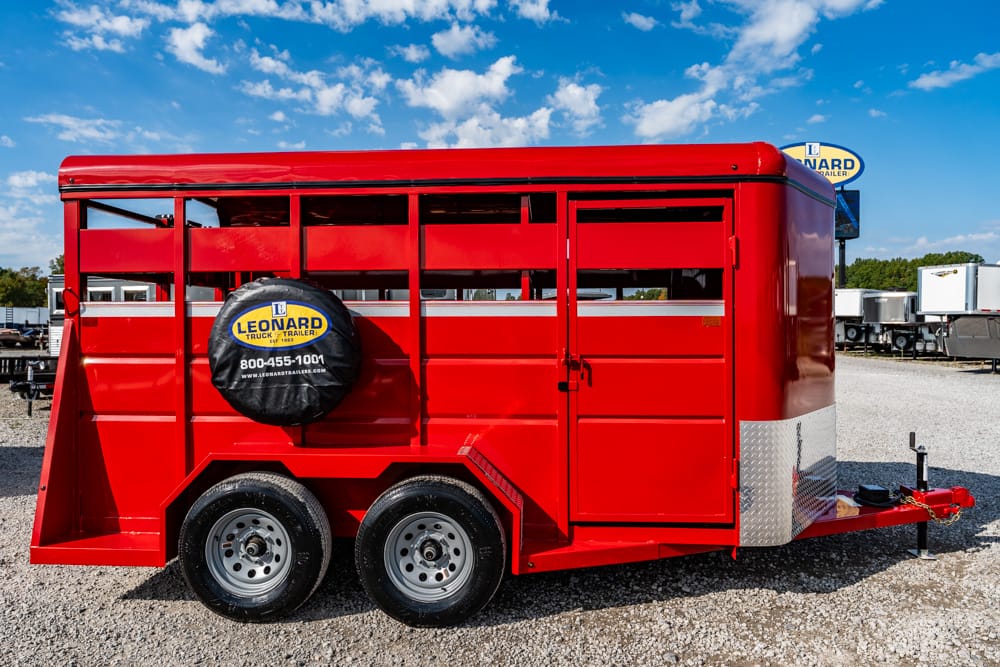 Red livestock trailer with a Leonard branding spare tire cover, parked on a gravel lot under a blue sky.