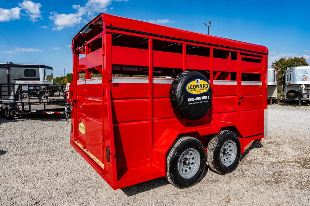 Red, two-axle livestock trailer with a mounted spare tire on the back, parked on a gravel lot with other trailers nearby.