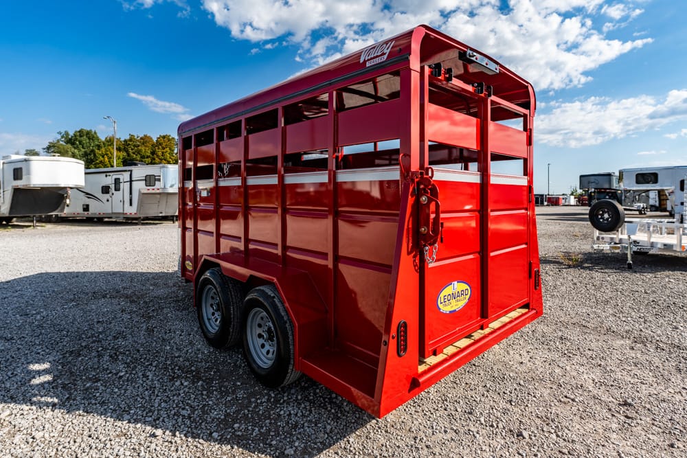 Red two-axle livestock trailer viewed from the rear-right, parked on a gravel lot with other trailers nearby.