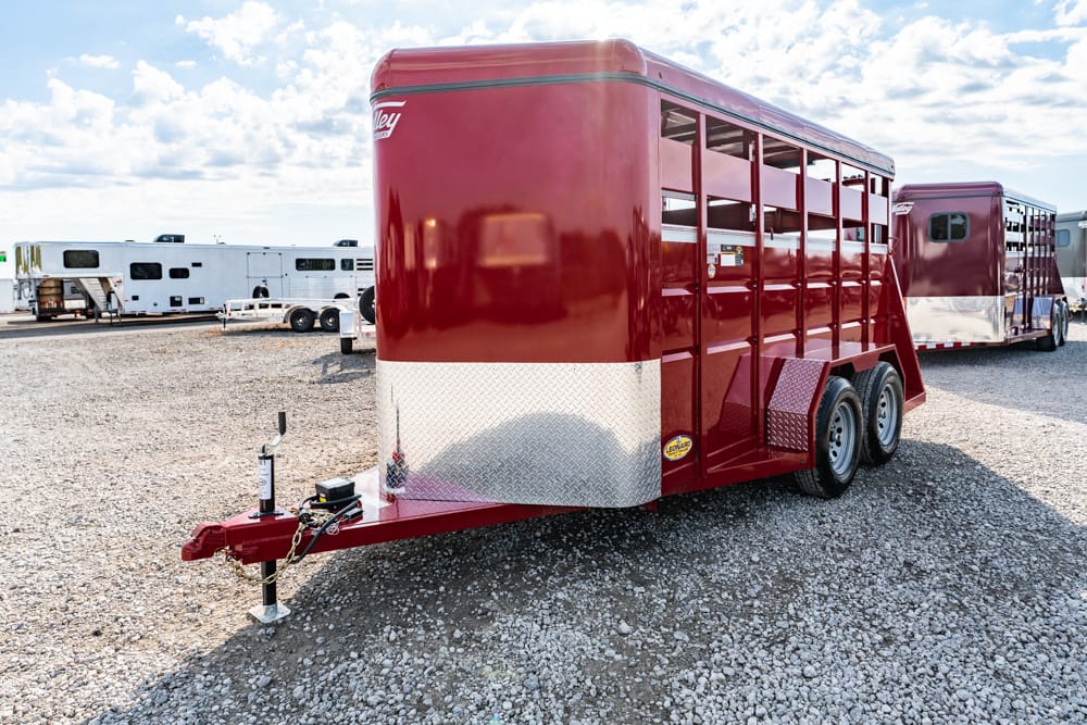Red livestock trailer with diamond-plate front parked on a gravel lot near other trailers in a yard.