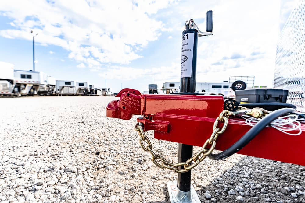 Close-up of a red trailer hitch with a gold safety chain in a gravel lot; RV trailers are visible in the background.