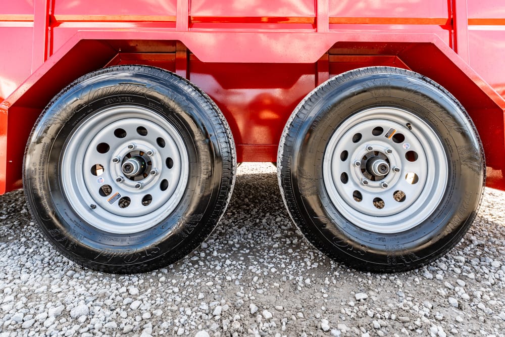 Two silver trailer wheels side by side under a red trailer on a gravel surface.