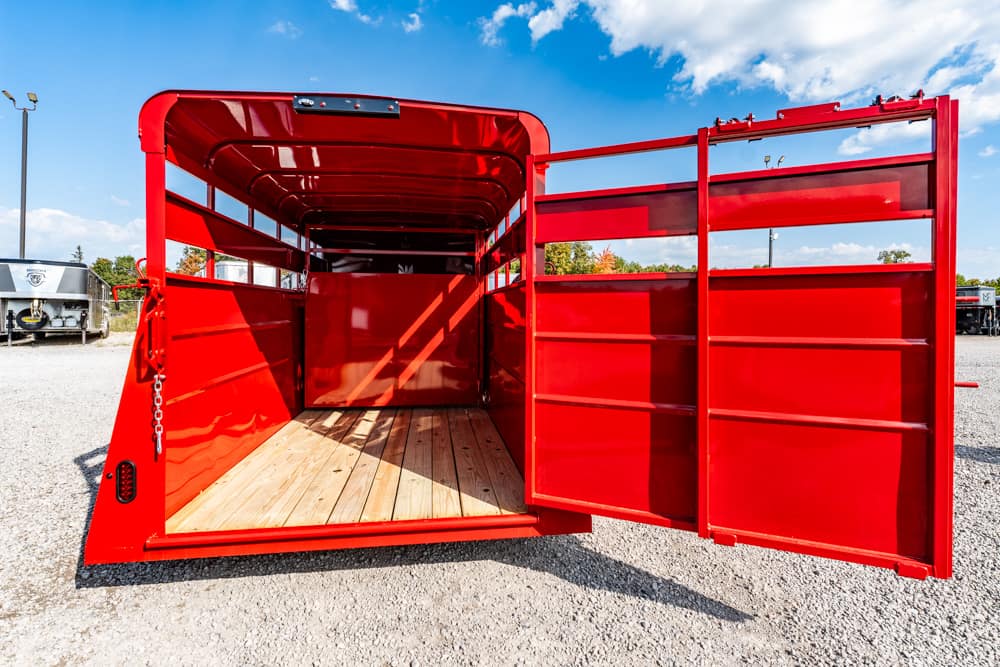 Red livestock trailer with rear double doors open, showing a wooden floor and empty stalls on a gravel lot under a blue sky.