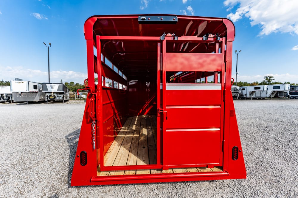 Open red livestock trailer with wooden floor, empty interior visible through rear doors, parked on a gravel lot under a blue sky