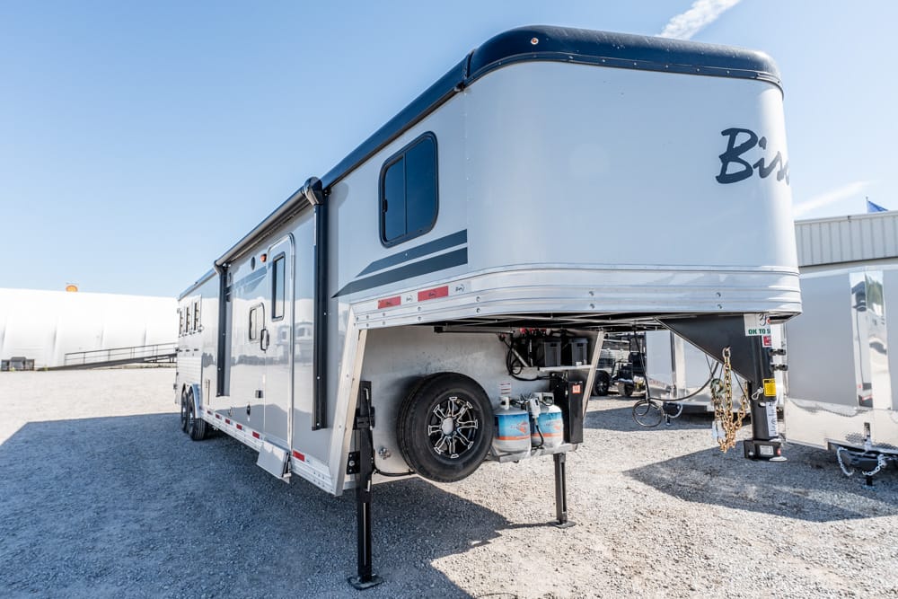 White and gray fifth-wheel trailer parked on gravel with stabilizer legs down, spare tire and propane tanks at the front.