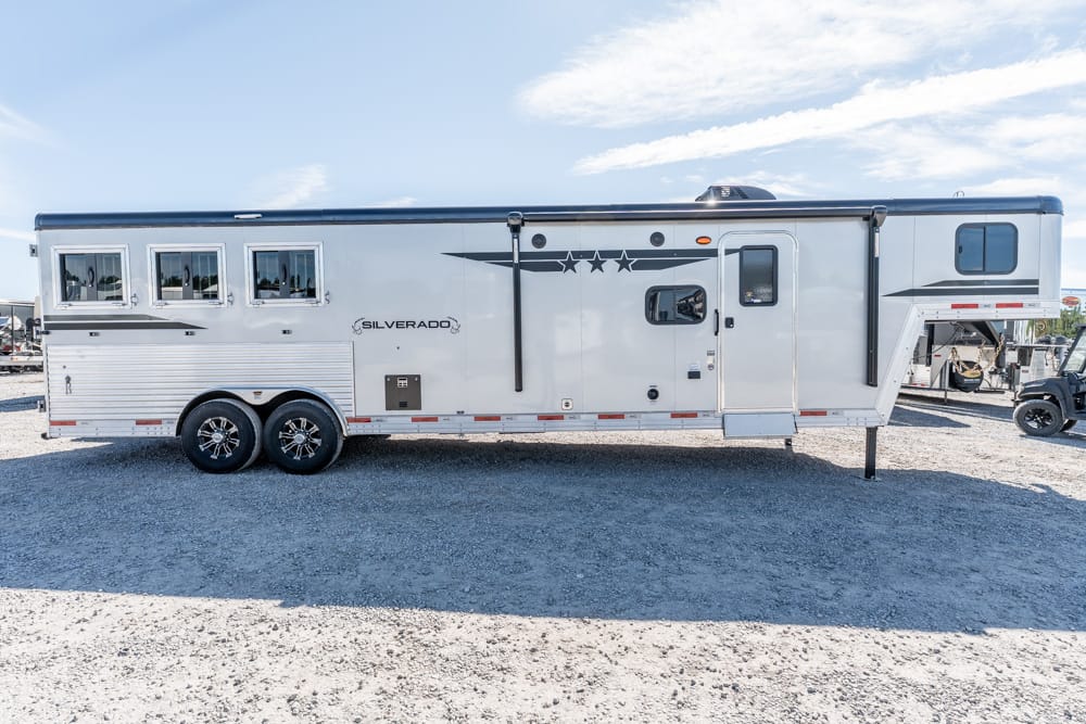White Silverado travel trailer with multiple windows, parked on a gravel lot under a blue sky.