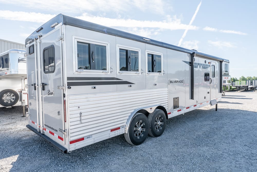 Long silver enclosed travel trailer with multiple windows, parked on gravel, two-axle wheels visible on the side