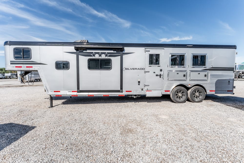 Long silver double-axle horse trailer with multiple windows and storage compartments, parked on a gravel lot.