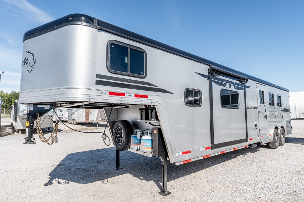 Silver gooseneck horse trailer parked on gravel, with propane tanks and spare tire visible under the body.