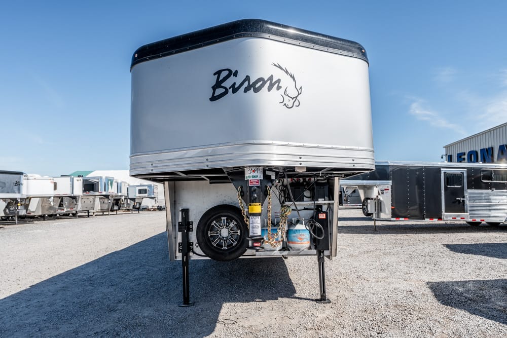 Front cap of a silver Bison gooseneck trailer raised on landing gear, with a spare tire, chains, and a propane tank visible in a gravel trailer lot.