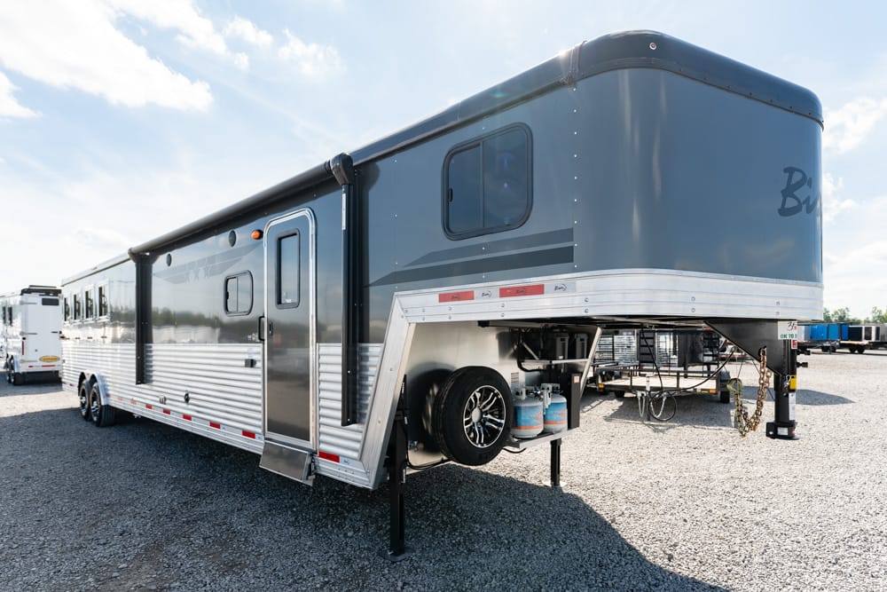 Silver fifth-wheel RV trailer on a gravel lot, with entry door, windows, and dual axles visible at the side.