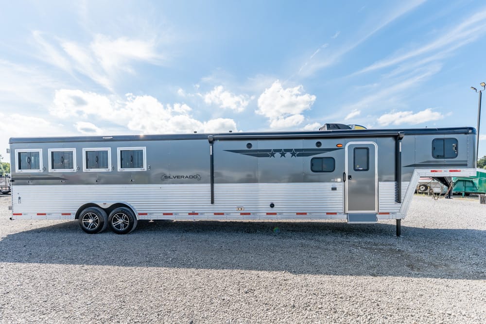 Silver and gray gooseneck horse trailer with four side windows and star decals on a gravel lot under a blue sky