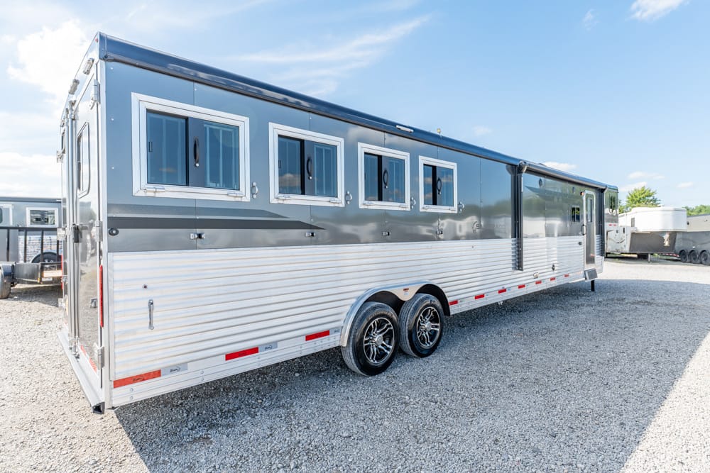 Silver aluminum horse trailer with four side windows and dual-axle wheels on a gravel lot.