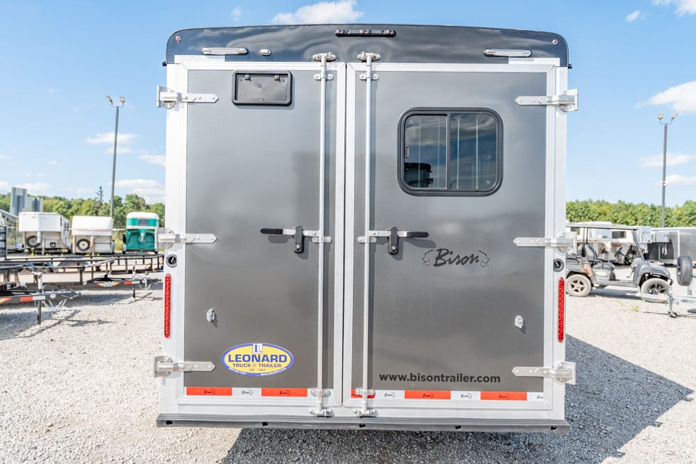 Rear view of a gray Bison horse trailer with rear window, metal latches, and brand logos in a dealership lot
