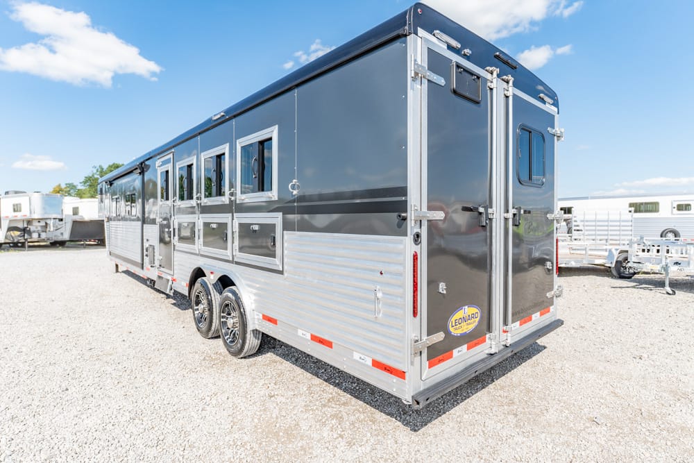 Long two-axle black-and-silver horse trailer with rear doors and multiple side windows on a gravel lot.