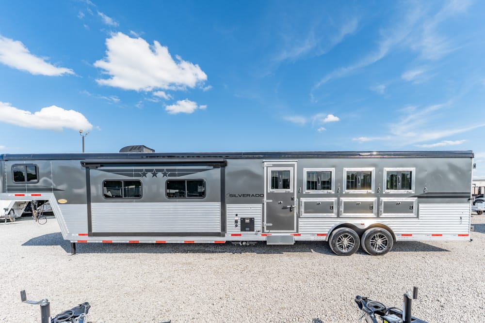 Long silver horse trailer with multiple horse stalls and windows parked on a gravel lot under a blue sky.