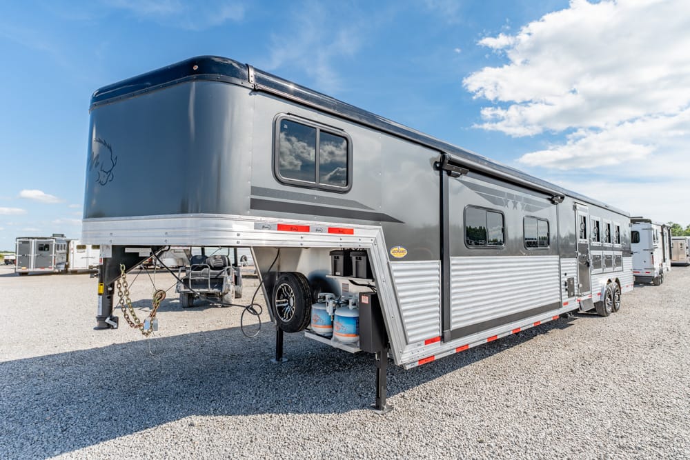 Grey gooseneck horse trailer with living quarters, propane tanks, spare tire, and awning on a gravel lot