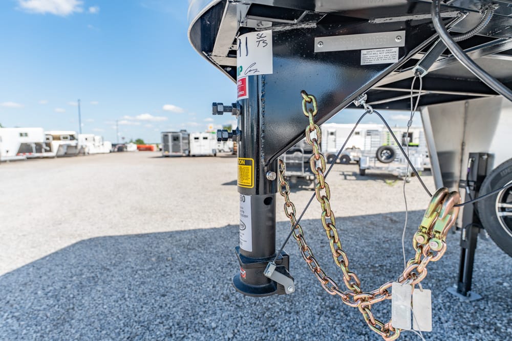 Close-up of a boat trailer hitch with safety chains, lock, and jack on a gravel lot, with trailers in the background