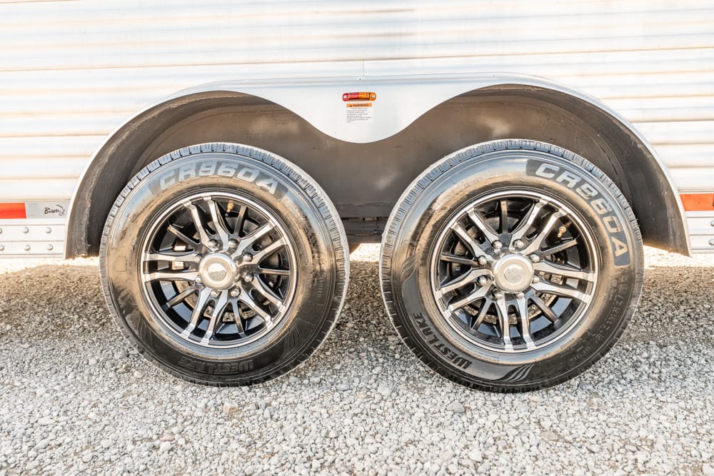 Dual-axle trailer with two black alloy wheels and tires on a gravel surface under a curved fender.