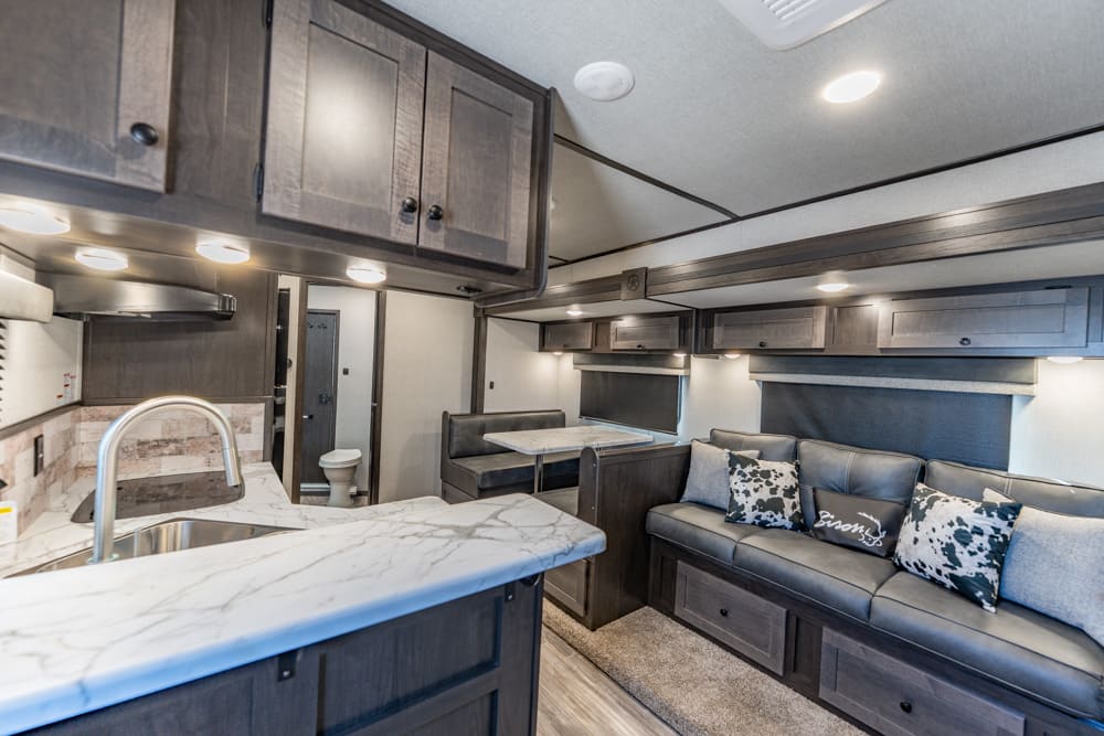 Interior of a modern RV: marble kitchen island with sink, dark cabinets above and below, and a gray sofa with patterned cushions in the living area.