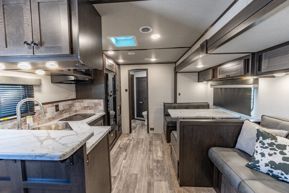 Interior of a modern RV: compact kitchen with marble countertops, sink, dark wood cabinets, and a dinette opposite a cushioned seating area.