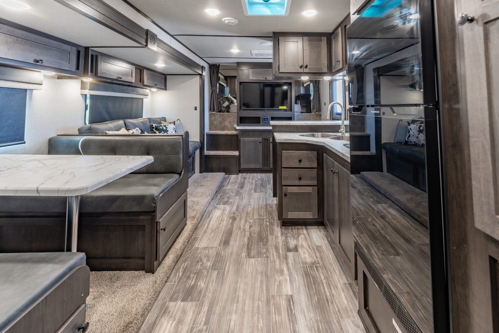 Interior of a modern travel trailer showing a kitchen with island, dinette, and sofa seating area with dark wood cabinets and marble countertops.