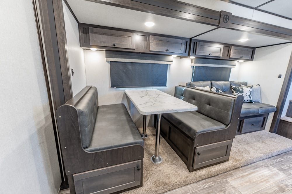 RV dining area with a marble-look table between two dark wood benches, overhead cabinets, and blue window shades.