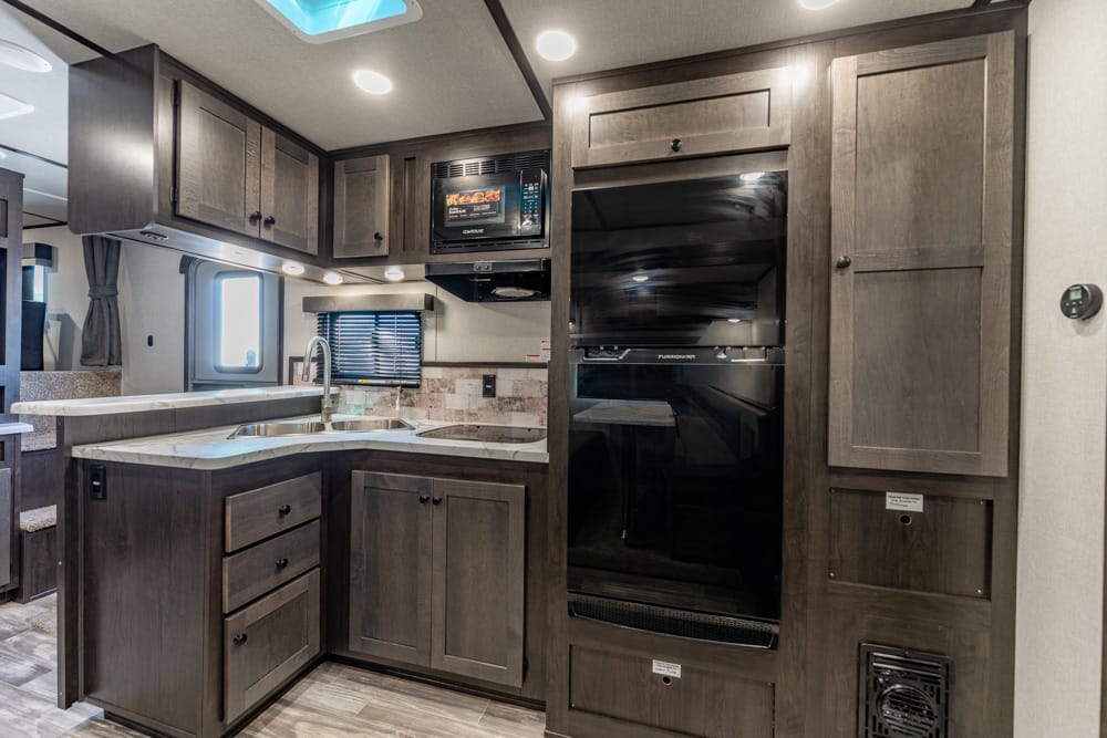 Interior of a compact RV kitchen with dark wood cabinets, a double sink, and a built-in black refrigerator.