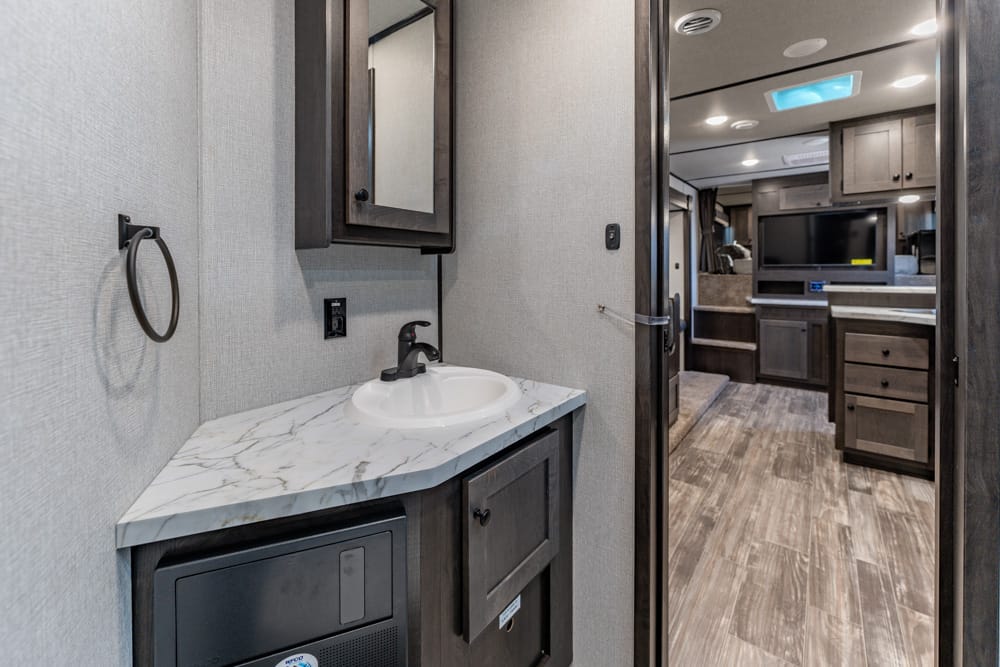 RV bathroom vanity with marble countertop, oval sink, dark wood cabinet, and towel ring; doorway to living area.