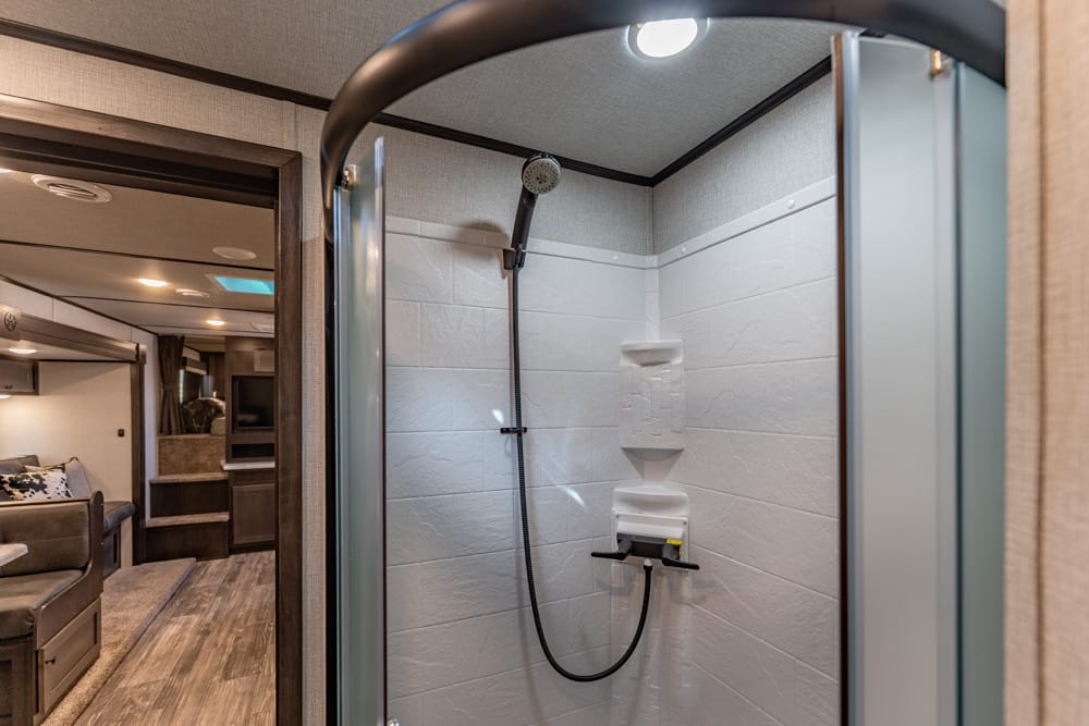 RV bathroom shower with white tile walls, built-in soap shelf, and a handheld black showerhead on a flexible hose.