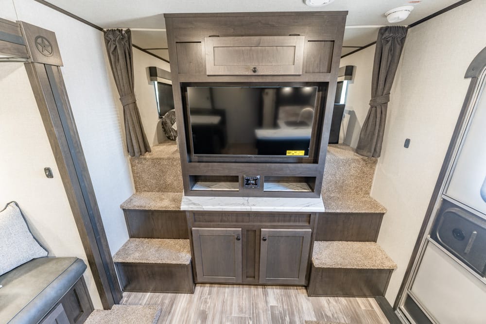 Dark wood RV entertainment center with a mounted TV, marble-like counter, and stepped carpeted benches on both sides.