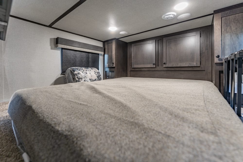 Compact RV bedroom with a large bed covered in beige blanket, dark wood cabinets along the wall, and ceiling lights above.
