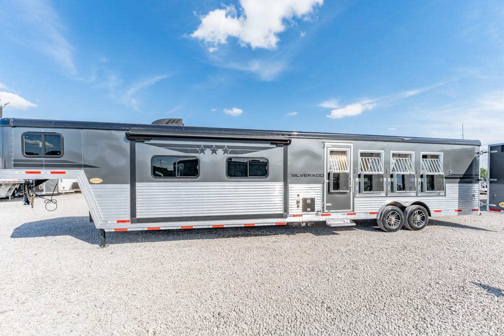 Silver horse trailer with multiple side windows and dual axles parked on a gravel lot under a clear blue sky.