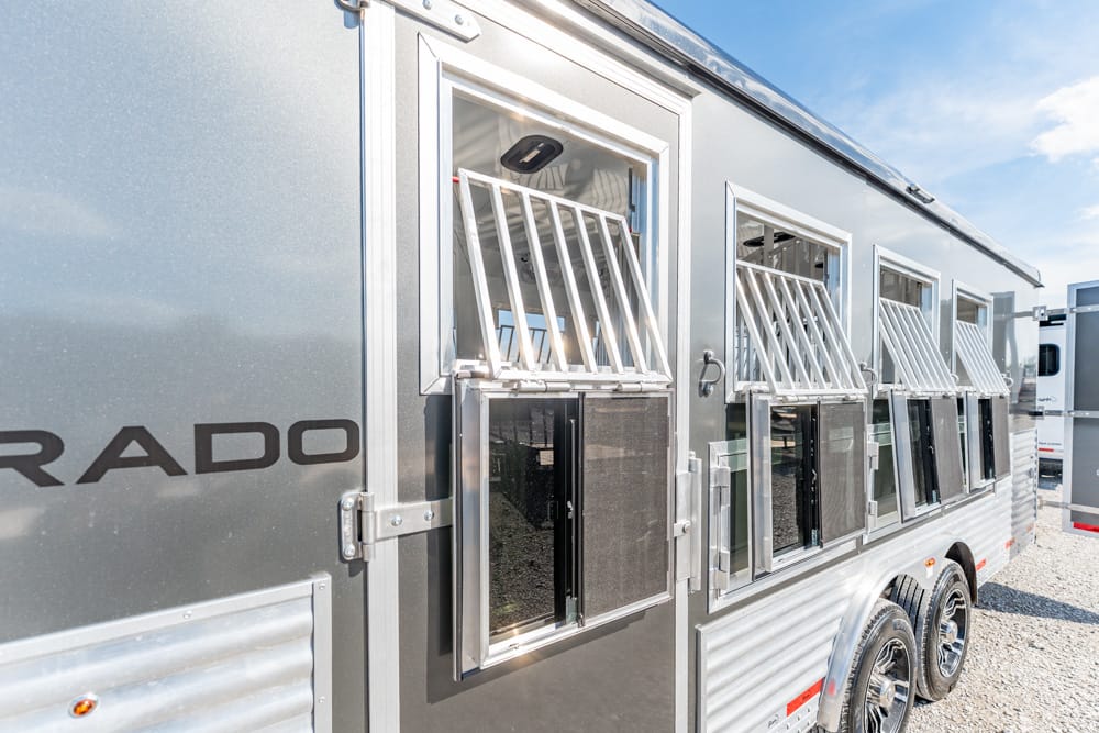 Silver horse trailer with open-barred side windows and dual-axle chrome wheels, parked on gravel under a blue sky.