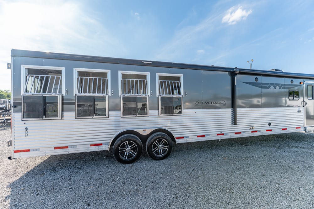 Long horse trailer with multiple windows and metal bars, parked on a gravel lot under a blue sky (brand: Silverado).