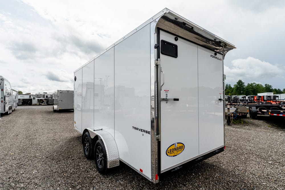 Rear three-quarters view of a white enclosed trailer with dual axles on a gravel lot; Leonard sticker on the back.