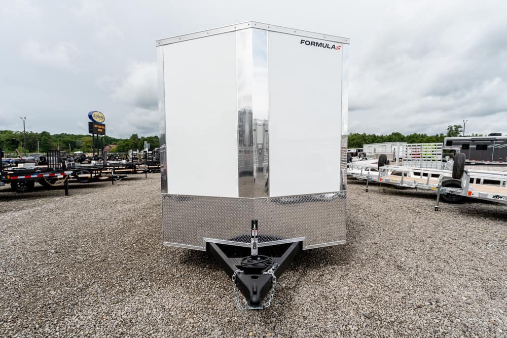 Front view of a silver-and-white enclosed cargo trailer with diamond plate at the front and a V-nose hitch, parked on a gravel lot with other trailers nearby.