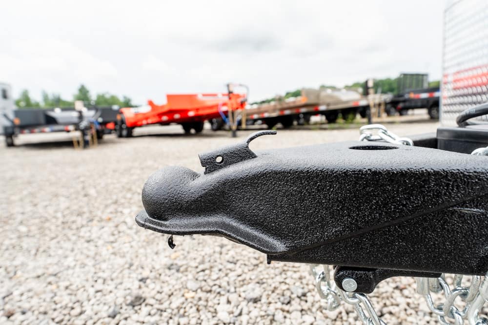 Close-up of a black textured trailer hitch coupler with safety chains, set on a gravel lot with trailers in the background.