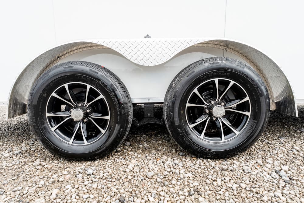 Dual trailer wheels with black-silver rims under a diamond-plate fender on a gravel surface