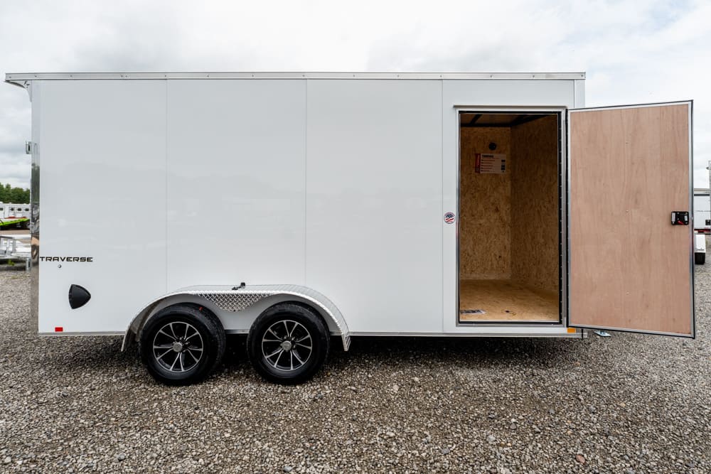 White enclosed cargo trailer with two axles and a wide rear door open to reveal a bare plywood interior on a gravel lot.