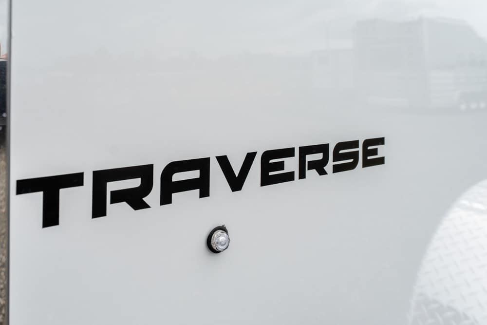 Close-up of the bold black TRAVERSE logo on a light gray trailer surface, with a small bolt beneath the text