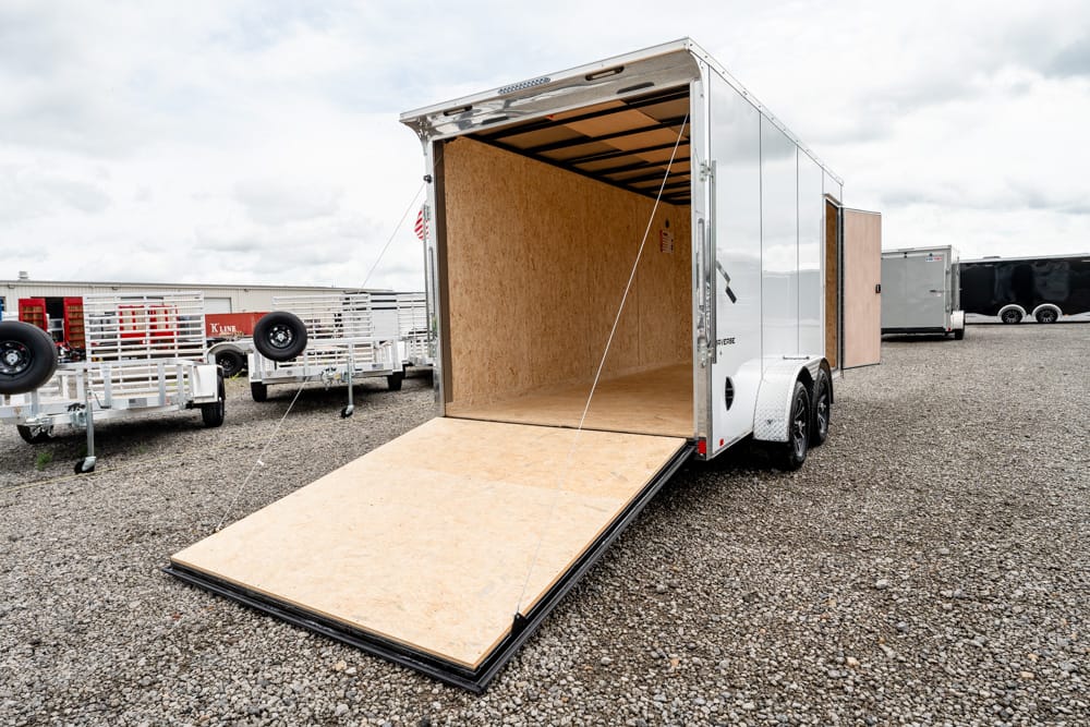 Enclosed two-axle trailer with ramp down, empty plywood interior, parked on gravel lot.