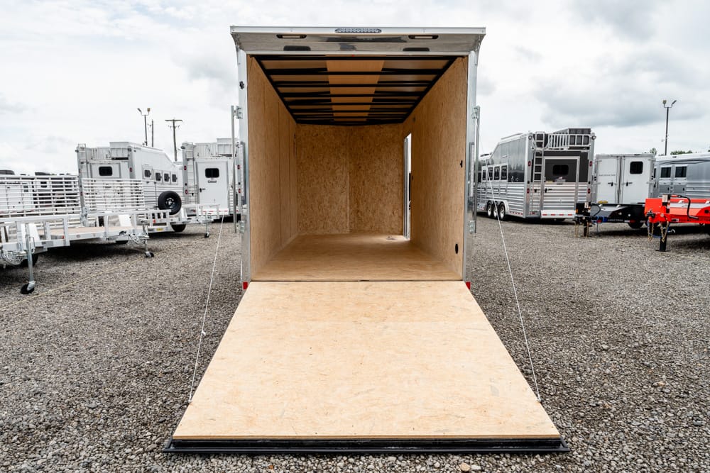 Open cargo trailer with a wooden ramp extended to the ground on a gravel lot, interior empty and plywood walls visible.