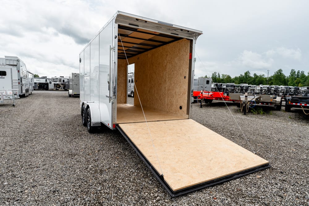 Enclosed white cargo trailer with an open ramp leading to a plywood interior, parked on a gravel lot with other trailers in the background.