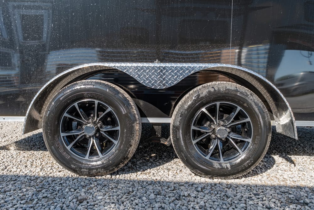 Dual-axle trailer wheel setup with black rims and diamond-plate fender on gravel surface.