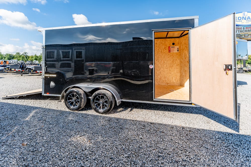 Black enclosed cargo trailer with an open door revealing a bare plywood interior on a gravel lot under a blue sky, sunlight gleaming on the surface.