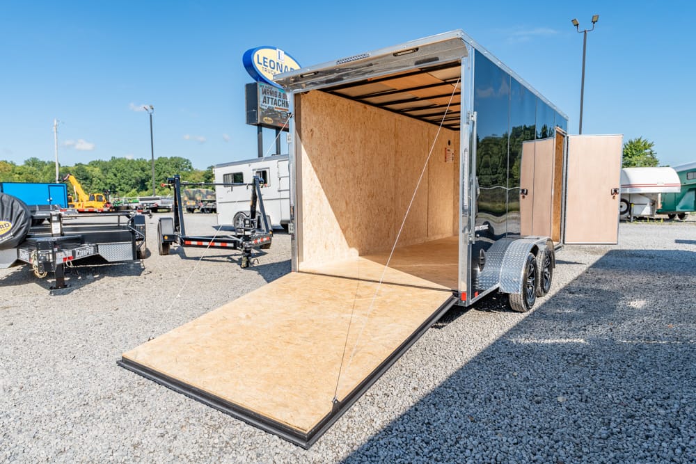 Enclosed cargo trailer with a lowered wooden ramp in a gravel lot, empty interior, and other trailers in the background.