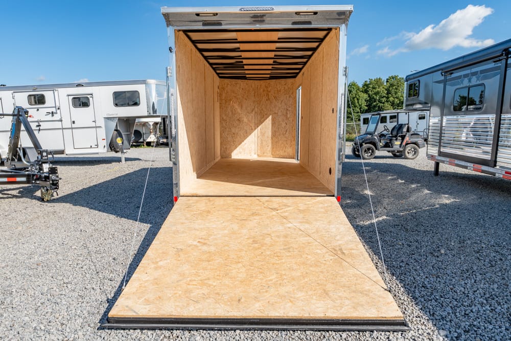 Open cargo trailer with ramp down, showing plywood interior, parked on gravel lot with other RVs nearby.